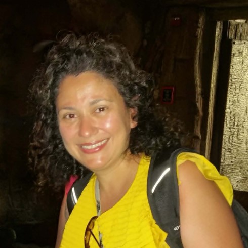 A woman with dark brown curly hair and brown eyes, wearing a yellow blouse.